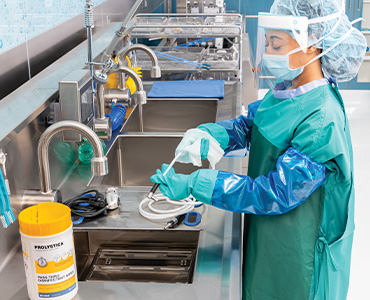 Sterile processing technician wearing sterile protective clothing, mask, and face shield cleans a medical instrument at a stainless steel sink using the PROLYSTICA Pass-Thru Disinfectant Wipe&nbsp;